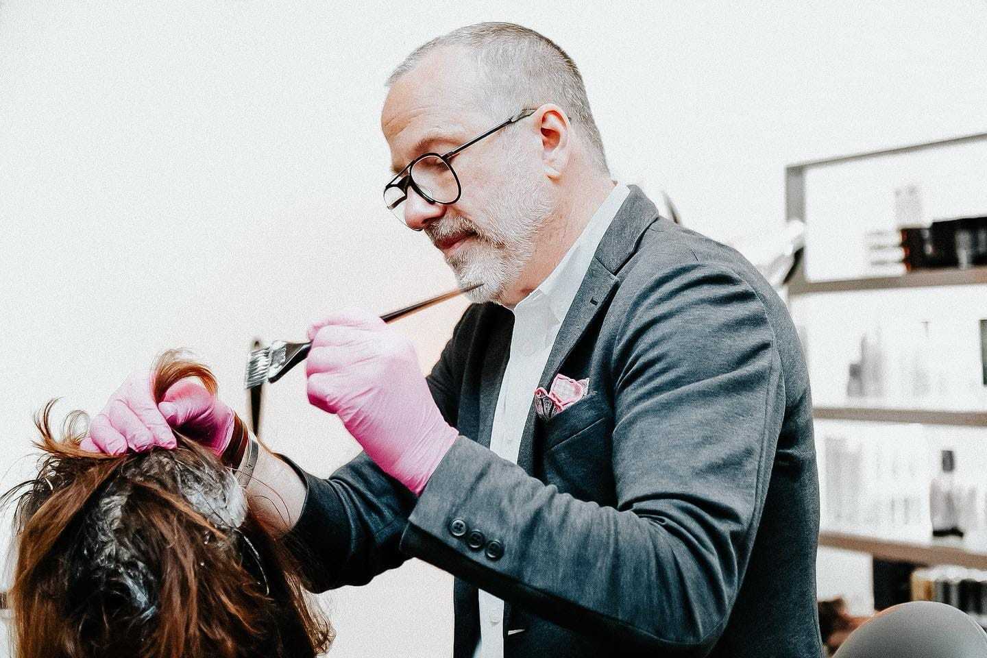 Man in glasses and pink gloves dyeing a person's hair in a modern salon.