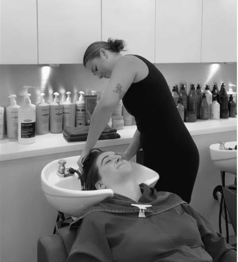 Hairdresser washing a client's hair in a salon, surrounded by shampoo bottles and hair products.