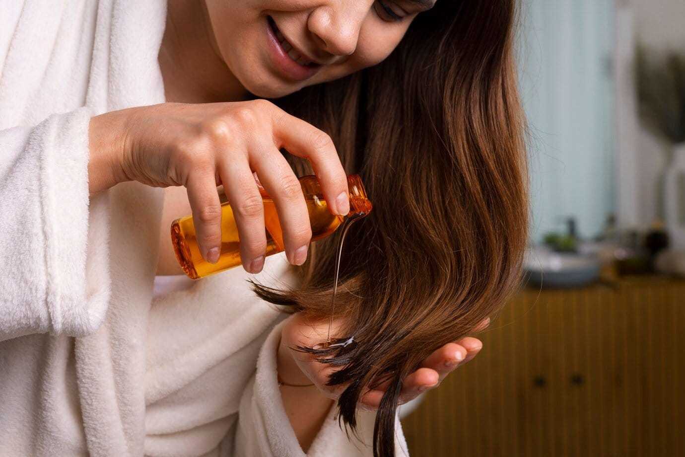 Woman applying hair oil to the ends of her long hair while wearing a white robe.