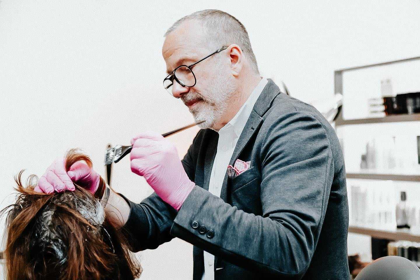 Man in glasses and pink gloves dyeing a person's hair in a modern salon.